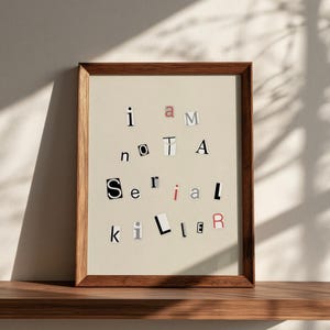 May include: A framed wall art print featuring the text 'i am not a Serial killer' composed of individual letters cut from various fonts and colors, resembling a ransom note style. The frame is made of medium-toned wood with a simple profile.