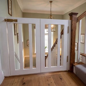 May include: A white wooden baby gate with vertical slats and a brass latch, installed at the top of a staircase. The gate is hinged and appears to be closed, blocking access to the stairs. The surrounding walls are painted a light green.