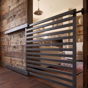 May include: A contemporary sliding metal gate with horizontal bars, mounted on a track. The gate is dark grey and contrasts with the warm wood paneling and flooring. The design provides a visual separation while maintaining an open feel.