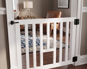 May include: A white wooden baby gate installed in a doorway. The gate has vertical slats and black hinges. The gate is closed, blocking the view into a living room with a rug and furniture.