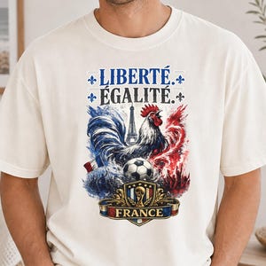 Puede incluir: Una camiseta de manga corta color blanco roto con un estampado gr&aacute;fico que incluye el texto LIBERT&Eacute; &Eacute;GALIT&Eacute;, un gallo, la Torre Eiffel, un bal&oacute;n de f&uacute;tbol y un escudo con la palabra FRANCE, en salpicaduras de estilo acuarela azul, blanco y rojo.