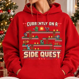 May include: A bright red hooded sweatshirt featuring a pixelated graphic of a video game level with platforms, ladders, gold coins, and small characters. The text reads "CURRENTLY ON A SIDE QUEST" in a blocky, retro-style font with small heart icons.