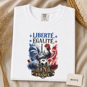 Puede incluir: Una camiseta blanca con un estampado gr&aacute;fico que incluye el texto LIBERT&Eacute; &Eacute;GALIT&Eacute;. El dise&ntilde;o muestra un gallo, la Torre Eiffel, un bal&oacute;n de f&uacute;tbol y un escudo con la palabra FRANCE, todo en salpicaduras de estilo acuarela azul, blanco y rojo.