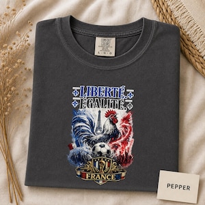 Puede incluir: Una camiseta gris carb&oacute;n con un estampado gr&aacute;fico que incluye el texto LIBERT&Eacute; &Eacute;GALIT&Eacute;, un gallo, la Torre Eiffel, un bal&oacute;n de f&uacute;tbol y un escudo con la palabra FRANCE. La camiseta tiene una etiqueta de la marca Comfort Colors en el cuello.
