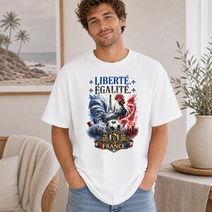 Puede incluir: Una camiseta blanca de manga corta con un estampado gr&aacute;fico de un gallo, un bal&oacute;n de f&uacute;tbol y la Torre Eiffel. El texto dice LIBERT&Eacute;, &Eacute;GALIT&Eacute; y FRANCE. El dise&ntilde;o incorpora colores azul, blanco y rojo con detalles de flor de lis.