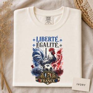 Puede incluir: Una camiseta de color marfil con un estampado gr&aacute;fico que incluye el texto LIBERT&Eacute; &Eacute;GALIT&Eacute; y FRANCE. El dise&ntilde;o muestra un gallo, la Torre Eiffel, un bal&oacute;n de f&uacute;tbol y un escudo dorado con un trofeo, todo en un estilo de acuarela azul, blanco y rojo.
