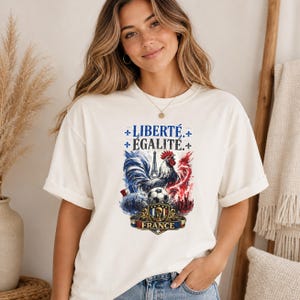 Puede incluir: Una camiseta de manga corta color blanco roto con un estampado gr&aacute;fico de un gallo, un bal&oacute;n de f&uacute;tbol y la Torre Eiffel. El texto dice LIBERT&Eacute; &Eacute;GALIT&Eacute; y FRANCE. El dise&ntilde;o incorpora colores azul, blanco y rojo con s&iacute;mbolos de flor de lis.