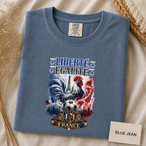 Puede incluir: Una camiseta de color azul jean con un estampado gr&aacute;fico que incluye el texto LIBERT&Eacute; &Eacute;GALIT&Eacute; y FRANCE. El dise&ntilde;o muestra un gallo, la Torre Eiffel, un bal&oacute;n de f&uacute;tbol y un escudo con detalles en acuarela de color rojo, blanco y azul.
