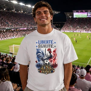 Puede incluir: Una camiseta blanca de manga corta con un estampado gr&aacute;fico que incluye el texto LIBERT&Eacute; &Eacute;GALIT&Eacute; y FRANCE. El dise&ntilde;o presenta un gallo, la Torre Eiffel, un bal&oacute;n de f&uacute;tbol y colores azul, blanco y rojo con detalles de flor de lis.