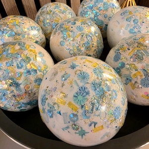 May include: A collection of eight spherical white ornaments decorated with an assortment of light blue and yellow floral-shaped sequins, glitter, and small rectangular confetti pieces embedded across the smooth, glossy surface of each round object.