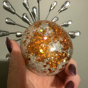 May include: A spherical ornament filled with clear liquid, white glitter, and large orange hexagonal glitter pieces. Several silver star-shaped sequins are suspended within the sphere. The ornament is held against a silver sunburst wall decor.