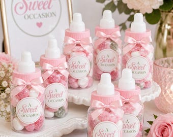 May include: A collection of baby bottle-shaped jars filled with sweets in pink, white, and green. Each jar is adorned with a pink bow and a label reading "A Sweet Occasion." The jars are displayed on a tiered serving dish, with a framed sign in the background.