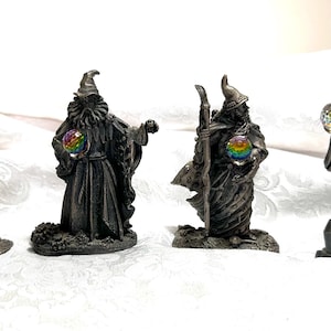 Könnte beinhalten: Vier Zinn-Zaubererfiguren mit detaillierten Roben, Hüten und Bärten. Zwei Zauberer halten leuchtende Kugeln, während sich ein anderer auf einen Stab stützt. Die Figuren stehen auf einzelnen Sockeln und zeigen ein Fantasy-Thema.