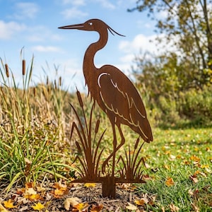 May include: A rust-colored metal garden stake featuring a silhouette of a standing heron bird surrounded by stylized cattail plants. The flat, cutout design displays a weathered, oxidized finish with detailed outlines of the bird's neck and wings.