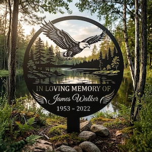 May include: A circular black metal memorial sign featuring a laser-cut design of a soaring eagle above a forest and lake landscape. The text reads IN LOVING MEMORY OF James Walker 1953 - 2022, with decorative wing accents at the bottom.
