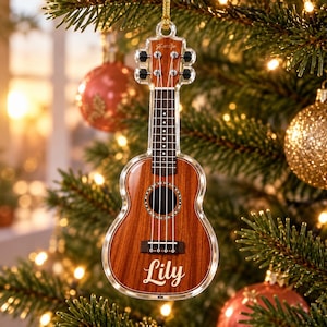 Könnte beinhalten: Ein durchsichtiger Acryl-Weihnachtsbaumschmuck in Form einer Miniatur-Ukulele mit Holzmaserung. Das Instrument hat vier Saiten, ein dunkles Griffbrett und den Namen Lily in weißer Schreibschrift auf dem Korpus.
