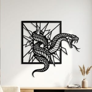 Può includere: Un'opera d'arte da parete in metallo nero che presenta una cornice quadrata con un effetto di vetro infranto. Un serpente dettagliato con la bocca aperta e le zanne visibili emerge dal centro della cornice, con il corpo che si estende.