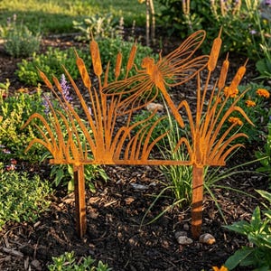 May include: A rusty metal garden stake featuring a dragonfly and cattails design. The dragonfly is in flight above the cattails and reeds. The metal has a weathered, orange-brown patina, and the stake is designed to be placed in a garden bed.