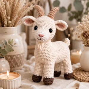 May include: A cream-colored crocheted goat toy featuring two light brown horns, dark brown hooves, black safety eyes, a small pink nose, and a short beard. The stuffed animal is crafted with visible yarn stitches and has a small tail.