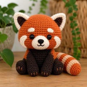 May include: A crocheted red panda plush toy featuring a rust-orange head with white ear tips and cheek patches, black eyes, a dark brown chest and limbs, and a long, striped tail with alternating bands of rust-orange and cream-colored yarn.