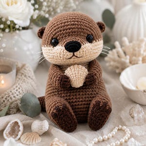 Può includere: Un peluche a forma di lontra realizzato all'uncinetto in marrone e beige con occhi in plastica nera e naso nero. La lontra ha baffi bianchi e tiene una piccola conchiglia beige lavorata all'uncinetto tra le zampe anteriori con punti testurizzati.