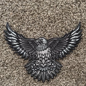 May include: An embroidered patch featuring a bald eagle in flight with wings fully spread. The design uses a monochromatic color palette of black, white, and various shades of gray to define the feathers, head, and talons of the bird.