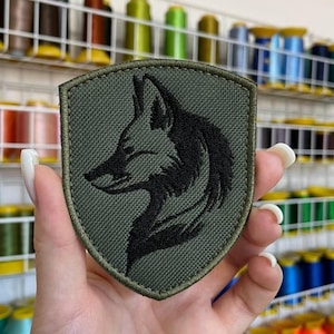 May include: An olive green embroidered patch with a black wolf head design. The patch is shield-shaped and has a textured appearance. The background shows spools of thread in various colors.