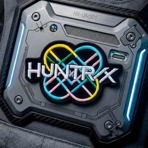 May include: Close-up of a gray, metallic device with the text "HX-UN001" at the top. A colorful patch with the word "HUNTRX" is affixed to the device. The patch features a multi-colored infinity symbol design. The device has blue LED lights.