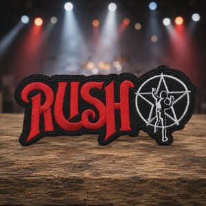 May include: An embroidered patch featuring the word RUSH in bold red lettering with a black border. To the right of the text is a white circular emblem containing a five-pointed star and a silhouette of a person standing with arms raised.