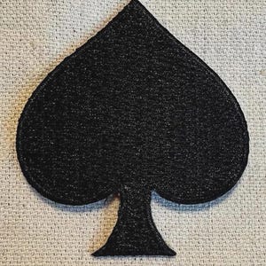 May include: A black spade symbol, a playing card suit, is centered on a textured, light gray background. The spade is a solid, dark color with a slightly glittery appearance.