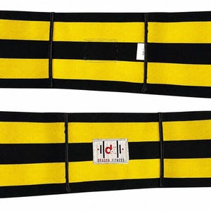 Op de afbeelding: Een rechthoekige fitnessband met horizontale gele en zwarte strepen. De voorzijde toont een wit rechthoekig label met de rode tekst "dk" en de zwarte tekst "DRAGON FITNESS" eronder. De achterzijde toont een klein wit stoffen label.