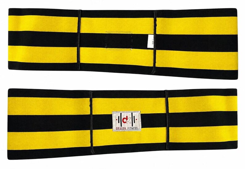 Op de afbeelding: Een rechthoekige fitnessband met horizontale gele en zwarte strepen. De voorzijde toont een wit rechthoekig label met de rode tekst "dk" en de zwarte tekst "DRAGON FITNESS" eronder. De achterzijde toont een klein wit stoffen label.