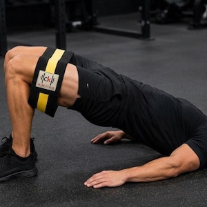 Op de afbeelding: Een zwarte weerstandsband met een gele middenstreep en een rechthoekige witte patch met de zwarte tekst "ck" en "DREAM FITNESS", geplaatst boven de knie&euml;n van een persoon die een glute bridge oefening uitvoert op een fitnessvloer.