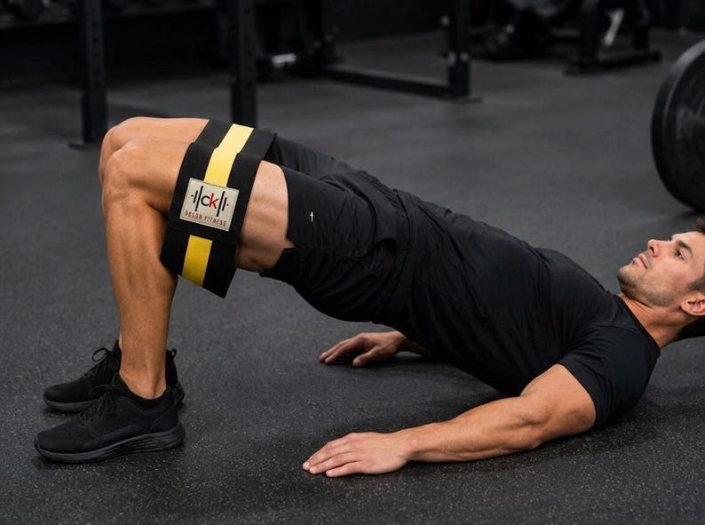 Op de afbeelding: Een zwarte weerstandsband met een gele middenstreep en een rechthoekige witte patch met de zwarte tekst "ck" en "DREAM FITNESS", geplaatst boven de knie&euml;n van een persoon die een glute bridge oefening uitvoert op een fitnessvloer.