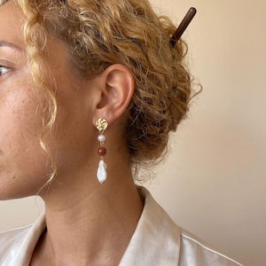 May include: Gold drop earrings with a textured disc, a white pearl, a reddish-brown bead, and a large, irregular white pearl. The earrings are designed to dangle, creating an elegant look. These earrings are a stylish accessory, suitable for various occasions.