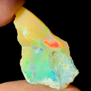 Puede incluir: Una piedra preciosa de ópalo en bruto con un color base amarillo cremoso. La piedra muestra destellos de rojo, verde, azul y naranja. El ópalo está sostenido por un dedo sobre un fondo negro, mostrando su forma natural y en bruto.