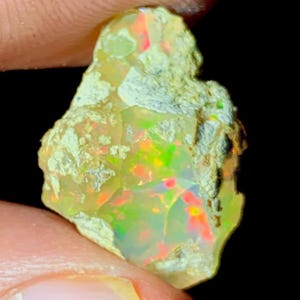 Puede incluir: Una piedra preciosa de ópalo en bruto con una vibrante exhibición de colores. La piedra presenta destellos de rojo, verde y amarillo dentro de una base translúcida y lechosa. El ópalo se sostiene sobre un fondo oscuro, resaltando su textura natural e iridiscencia.
