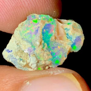 Op de afbeelding: Een ruwe, ongeslepen opaal edelsteen met een levendige kleurenpracht. De opaal vertoont flitsen van groen, blauw en oranje in een beige matrix. De steen heeft een natuurlijke, onregelmatige vorm.