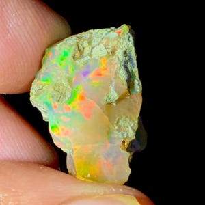 Puede incluir: Una piedra preciosa de ópalo en bruto con colores vibrantes. El ópalo muestra destellos de verde, naranja y púrpura dentro de una base blanca cremosa. La piedra tiene una superficie texturizada e irregular, y está sujeta por un dedo.