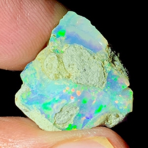 Puede incluir: Una piedra preciosa de ópalo en bruto con una vibrante exhibición de colores. La piedra presenta destellos de verde, azul y naranja sobre un fondo pálido y texturizado. El ópalo se sostiene entre los dedos, mostrando su forma natural e irregular.