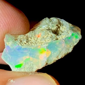 Op de afbeelding: Een close-up van een ruwe opaal edelsteen. De opaal toont een kleurenspel met flitsen van groen, oranje en blauw in een doorschijnende, melkachtige witte basis. De steen wordt tussen vingers gehouden tegen een zwarte achtergrond.