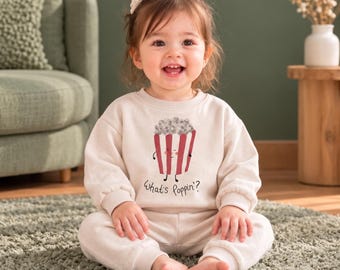 Puede incluir: Un conjunto de sudadera beige de manga larga y pantalón a juego con un gráfico de un cubo de palomitas de rayas rojas y blancas con una cara sonriente y el texto "What's poppin?" impreso en letra negra en la parte delantera.