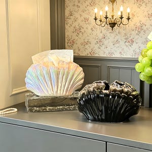 May include: Two decorative shell-shaped items. One is a pearlescent tissue box holder with tissues, and the other is a glossy black vase. Both are on a gray surface, with a chandelier and floral wallpaper in the background.