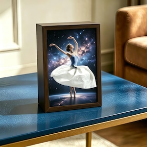 May include: A framed print of a ballerina in a blue leotard and white tutu, set against a starry, cosmic backdrop. The dark wood frame rests on a blue table with gold legs, creating a visual contrast. The frame is approximately 25 cm in height.