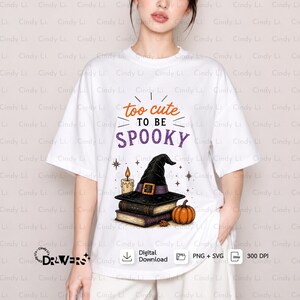 Puede incluir: Una camiseta blanca de manga corta con un estampado gráfico que incluye el texto "too cute TO BE SPOOKY" en naranja y morado. El diseño muestra un sombrero de bruja negro sobre una pila de libros, una vela encendida y una calabaza naranja.