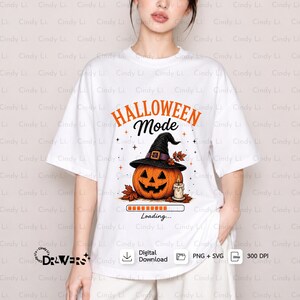 Puede incluir: Una camiseta blanca de manga corta con un gráfico de una calabaza con un sombrero de bruja negro, una vela encendida y hojas de otoño. El texto dice HALLOWEEN Mode y Loading... con una barra de carga debajo de la ilustración de la calabaza.