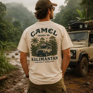 Può includere: Una t-shirt color crema con una grafica sul retro che riporta il testo CAMEL TROPHY '96 KALIMANTAN RAINFOREST EXPEDITION. Il disegno raffigura un veicolo fuoristrada circondato da palme e fogliame in inchiostro verde scuro e nero.