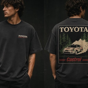 May include: Dark gray t-shirt with the Toyota logo on the front. The back of the shirt features a graphic of a rally car with the Toyota and Castrol logos. The design includes a forest scene and a vintage aesthetic.