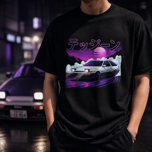 Könnte beinhalten: Schwarzes T-Shirt mit einer Grafik eines weißen und silbernen Autos, das auf einer lila und rosa Straße driftet, mit japanischem Text darüber. Das Design umfasst einen lila Sonnenuntergang und Berge. Das Shirt ist ein Freizeitartikel.