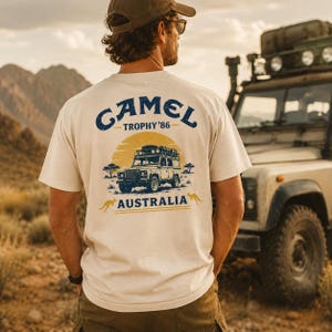 May include: A cream-coloured short-sleeve t-shirt featuring a graphic on the back with the text CAMEL TROPHY '86 AUSTRALIA. The design includes a yellow sun, a dark blue off-road vehicle, two small kangaroo silhouettes, and trees.